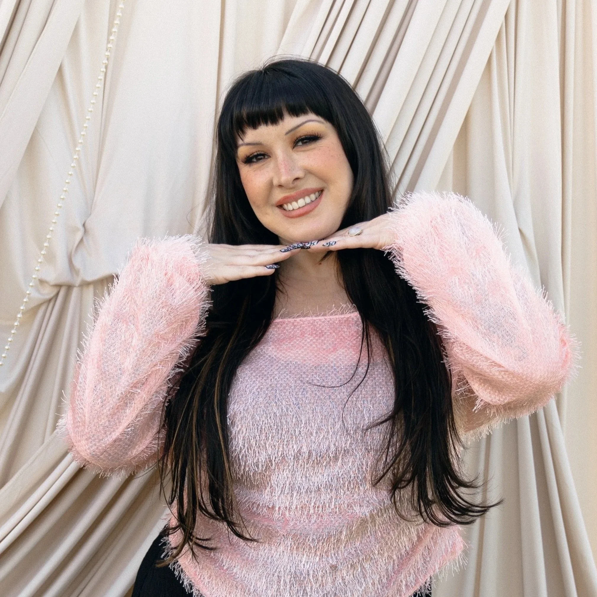 A woman with black hair and bangs smiling, posing with her hands under her chin, wearing a fluffy pink sweater, against a cream-colored curtain backdrop.