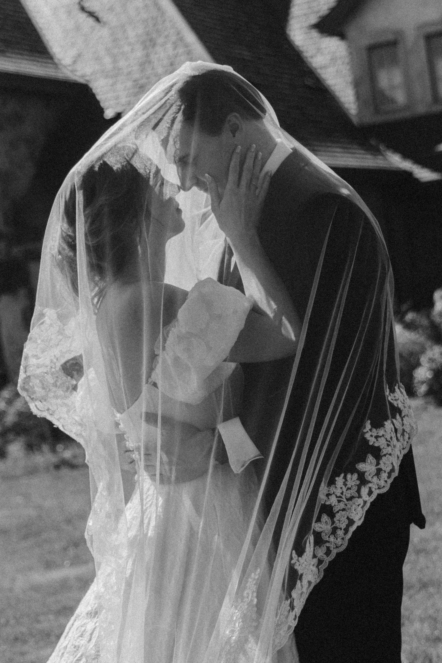 A black and white photo of a bride and groom sharing a moment, with the bride wearing a lace veil and dress, and the groom in a dark suit. They are close together, embracing and touching foreheads, in an outdoor setting.