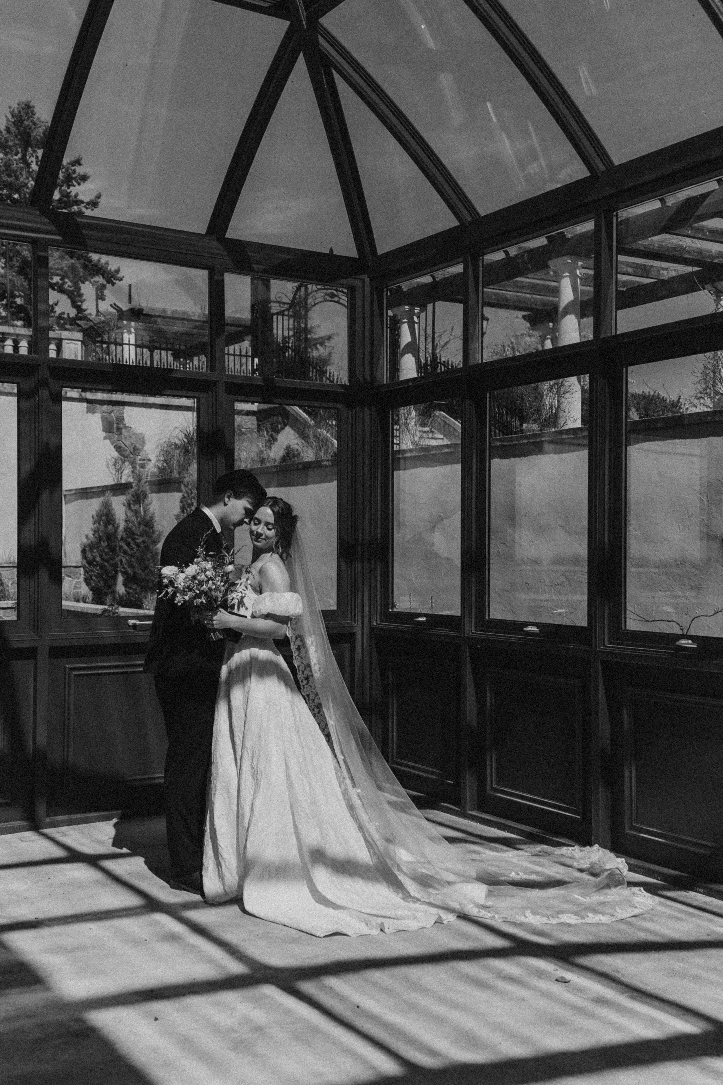 A Black and white photo of a wedding couple dancing inside a glass greenhouse, with the shadow of window frames cast on the wooden floor.