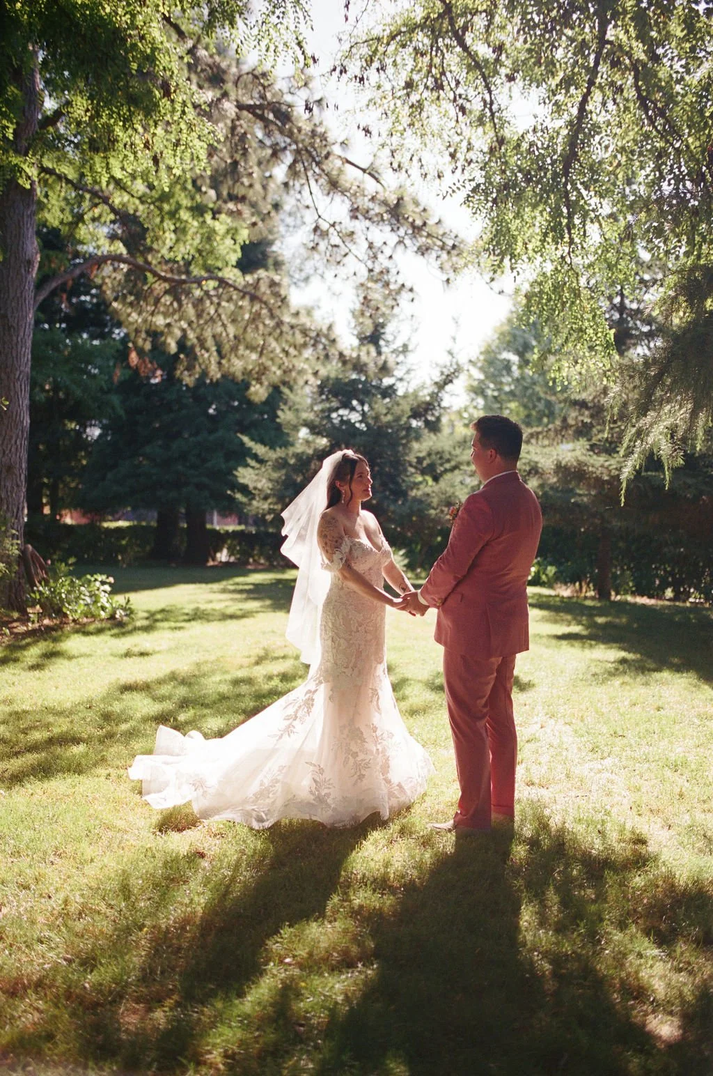 A bride and groom holding hands and facing each other outdoors under trees, during their wedding ceremony in the late afternoon.