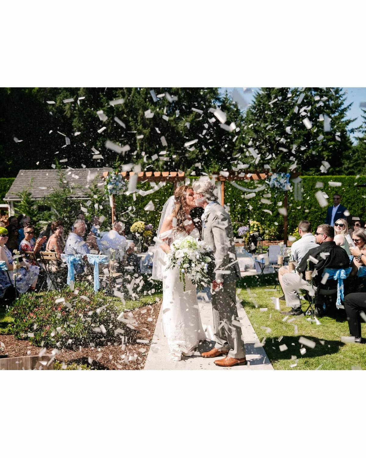 A wedding ceremony outdoors with a bride and groom sharing a kiss as confetti falls around them, surrounded by seated guests on a sunny day with trees and a decorated arch in the background.