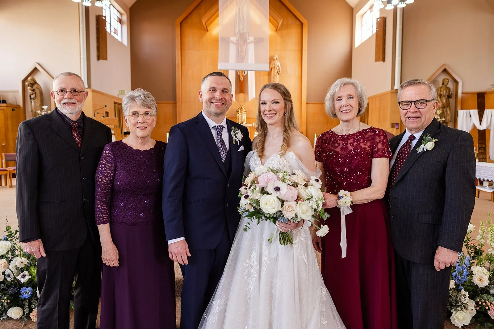 A wedding group photo inside a church features a bride and groom in the center, surrounded by four older adults, likely family members. The bride wears a white wedding dress and holds a bouquet, while the groom wears a suit with a boutonniere. The ot