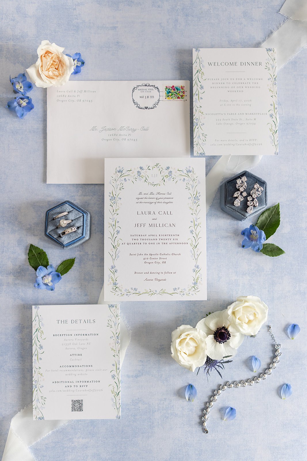 Wedding invitation suite with floral accents, jewelry, and flowers arranged on a light blue background, including an envelope, RSVP card, reception details card, and main invitation card.