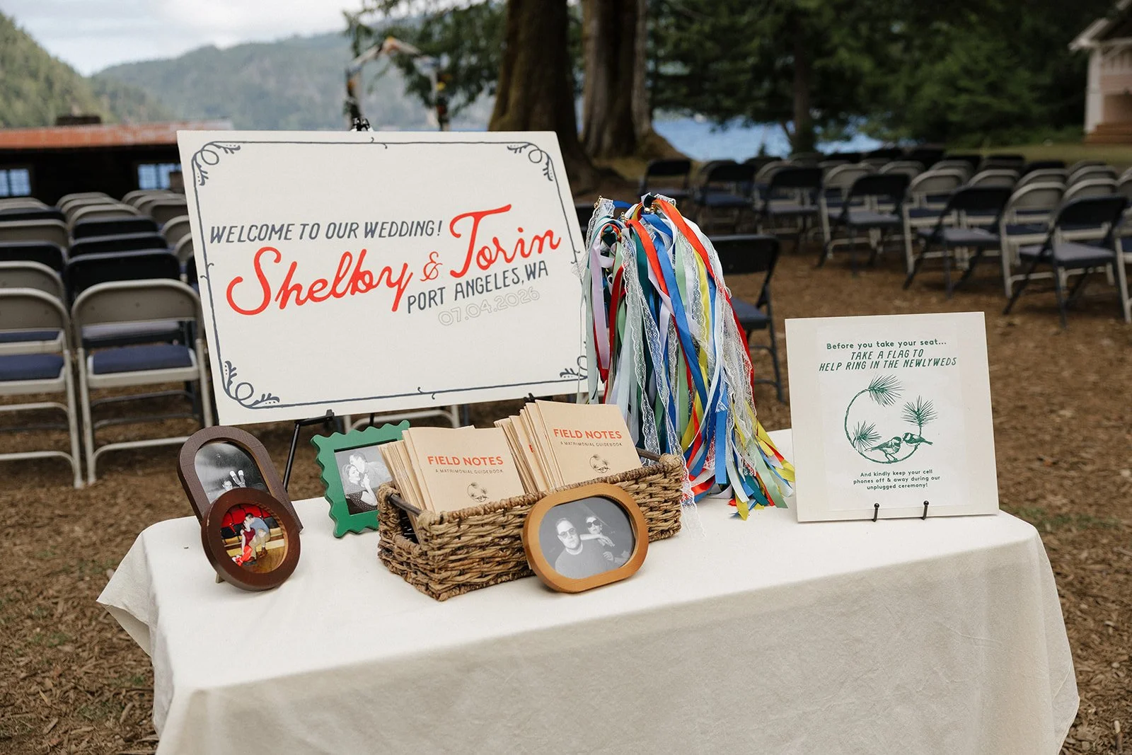 Wedding welcome table with framed photos, field notes, colorful ribbons, and a sign that says "Welcome to our wedding! Shelby & Torin Port Angeles, WA 07.04.208" in an outdoor setting with chairs and trees in the background.