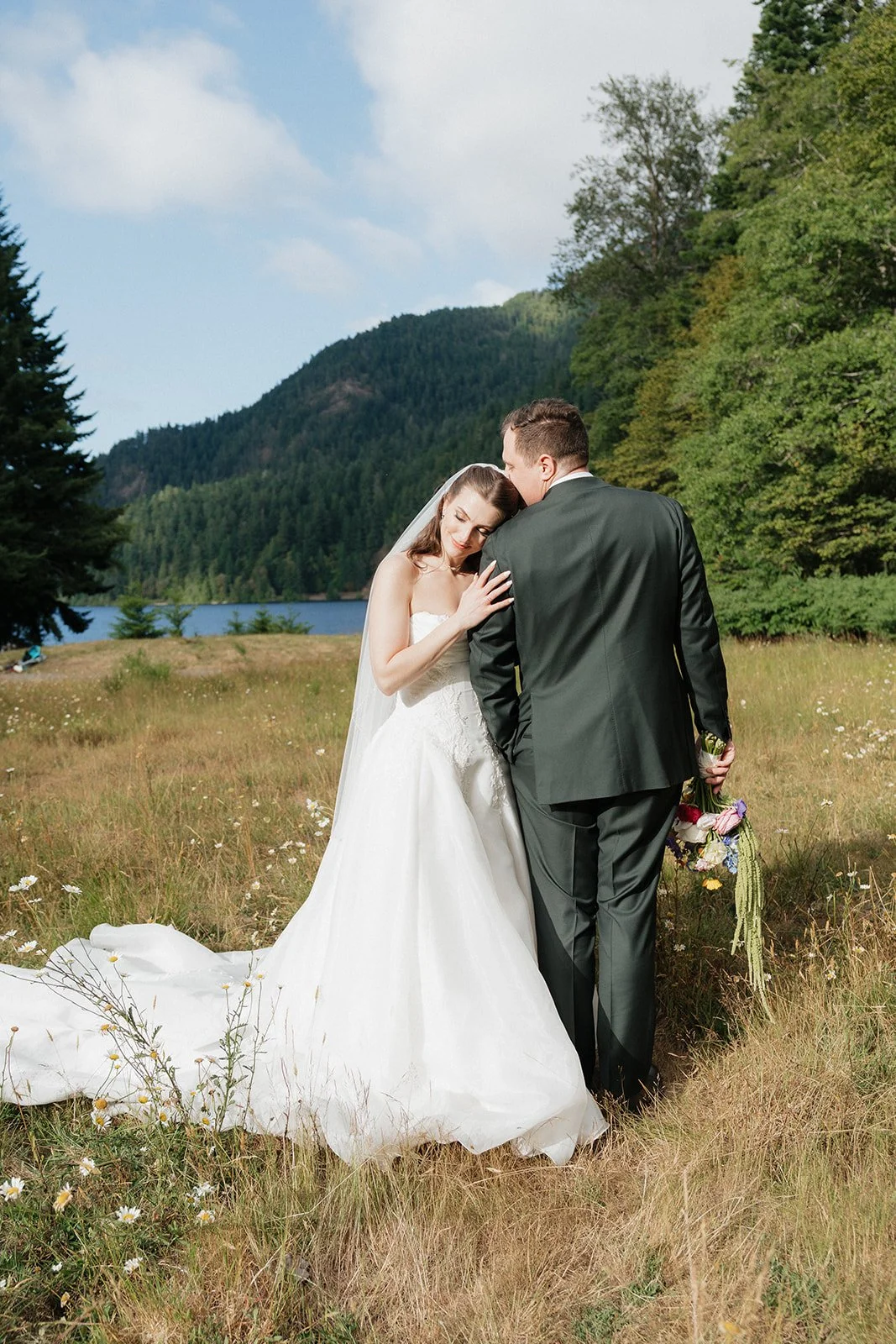 A bride and groom embracing in a grassy field with trees, mountains, and a lake in the background on a partly cloudy day.