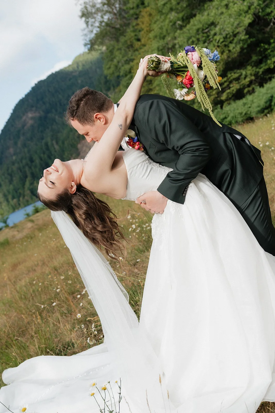 A wedding couple dancing outdoors, with the groom dipping the bride backwards. The groom is wearing a black suit and the bride a white wedding gown with a veil, holding a bouquet of flowers. Green hills and trees are in the background.