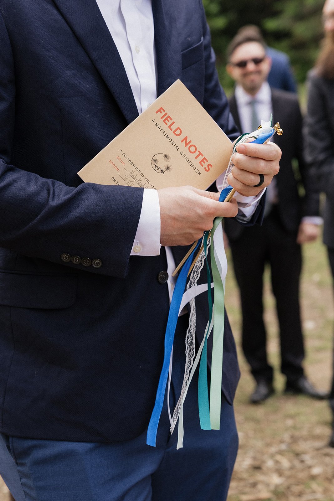 Man in a suit holding a booklet titled "Field Notes: A Matrimonial Guidebook" and wedding ribbons, with people in the background.