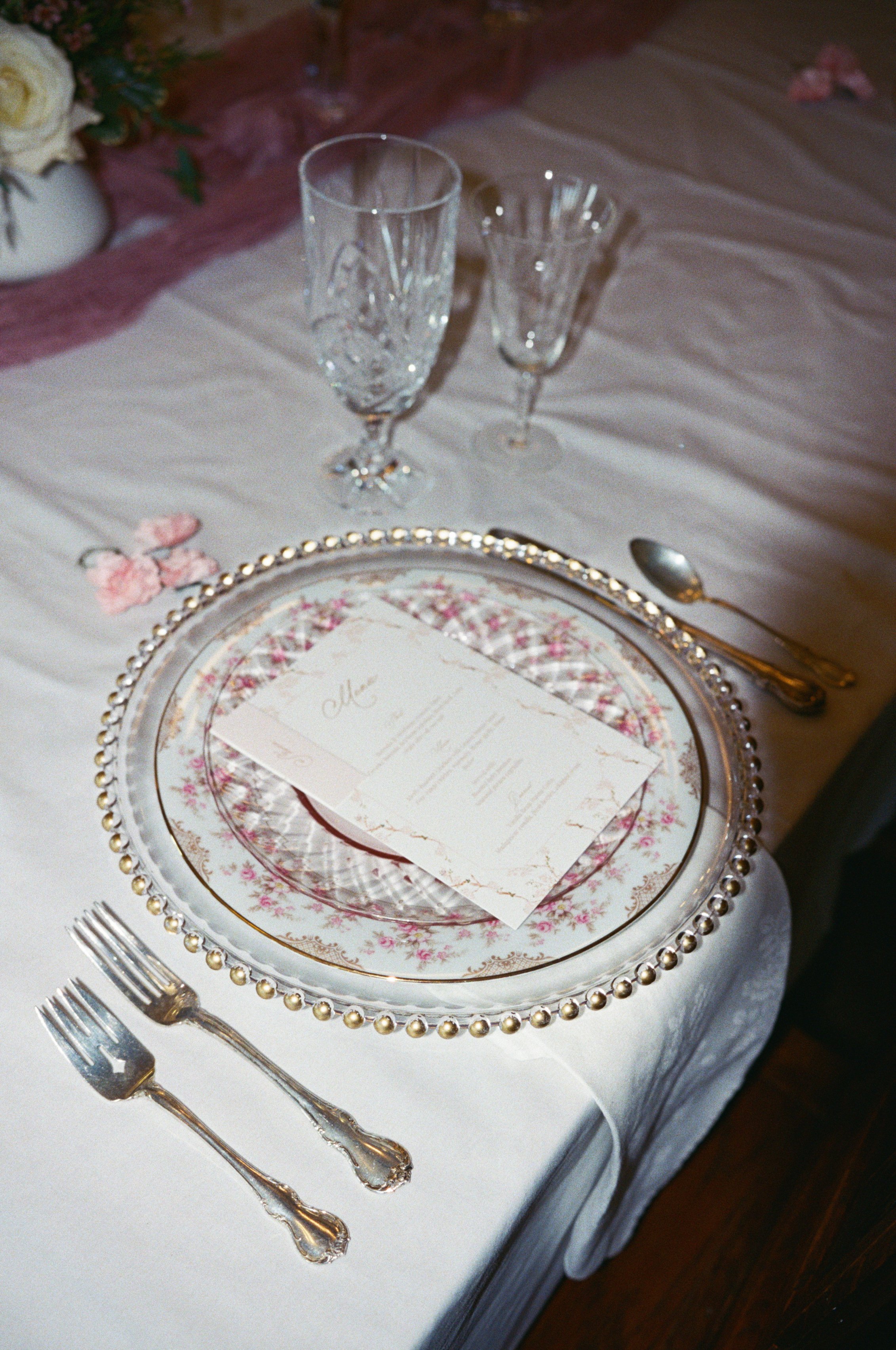 Elegant table setting with two crystal champagne flutes, a decorative plate with a floral design, and silverware on a white tablecloth, with pink and white flowers in the background.