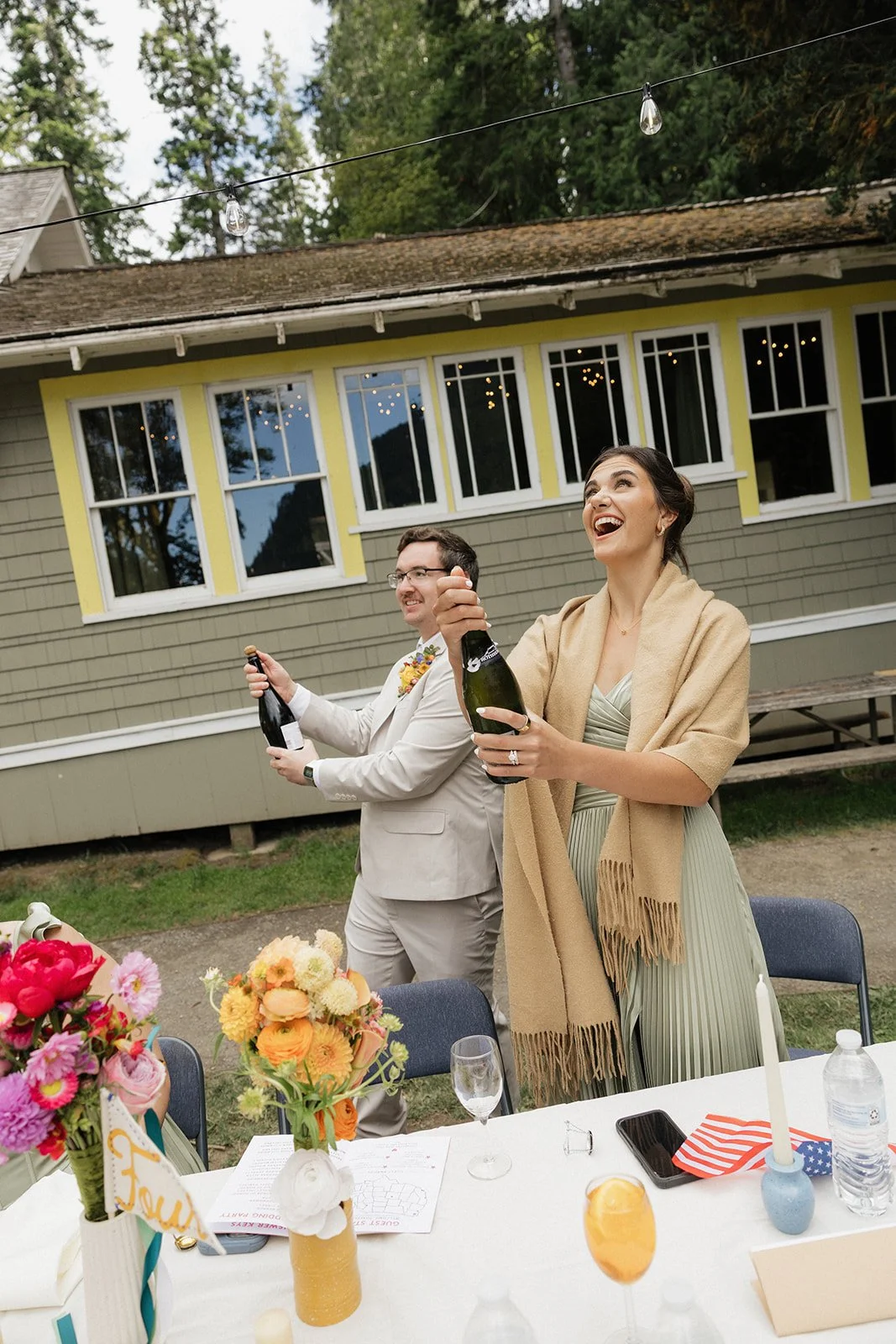 A woman and a man celebrating outdoors, holding bottles of champagne. The woman is wearing a beige shawl and a pleated dress, while the man is in a light suit with a boutonnière. They are standing behind a table decorated with flowers, candles, and d