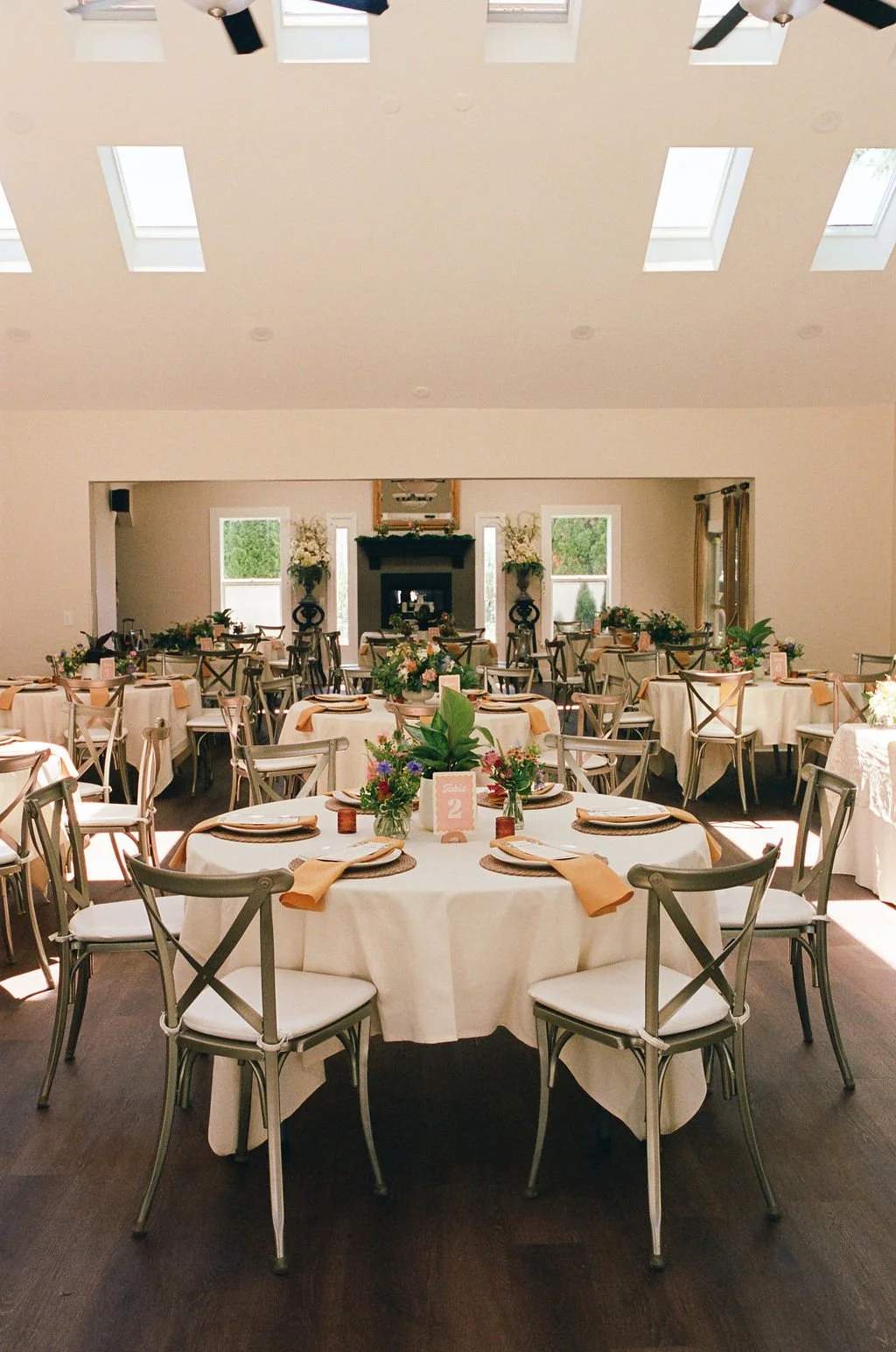 A spacious banquet hall decorated for a wedding reception with round tables covered in white tablecloths and beige napkins, floral centerpieces, wooden chairs, and a fireplace at the back flanked by vases with flowers. Natural light streams in throug