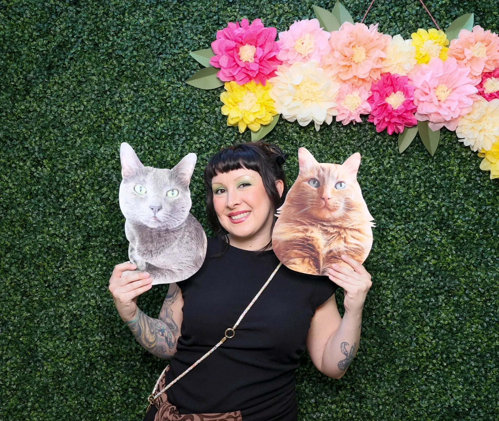 Woman with tattoos holding two cat photos in front of a green leafy wall decorated with pink, yellow, and white paper flowers.