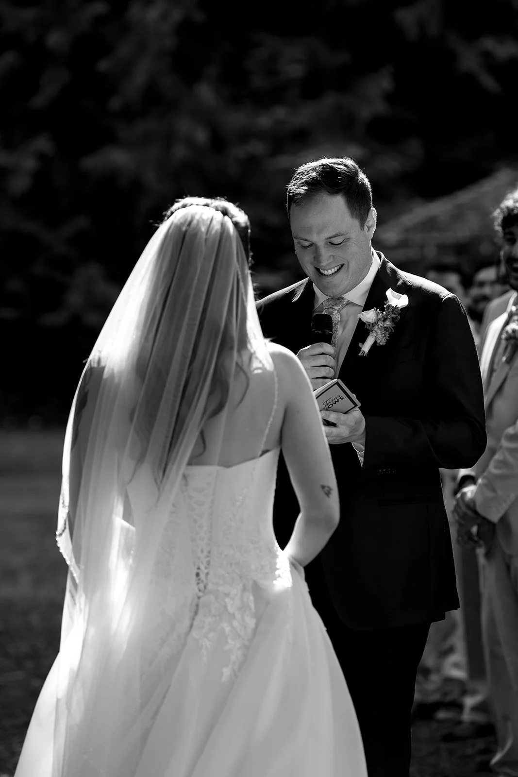 A black and white photo of a wedding ceremony outdoors, with a bride and groom exchanging vows. The groom is smiling and holding a microphone, dressed in a suit with a boutonniere. The bride is wearing a strapless wedding gown and veil, facing the gr