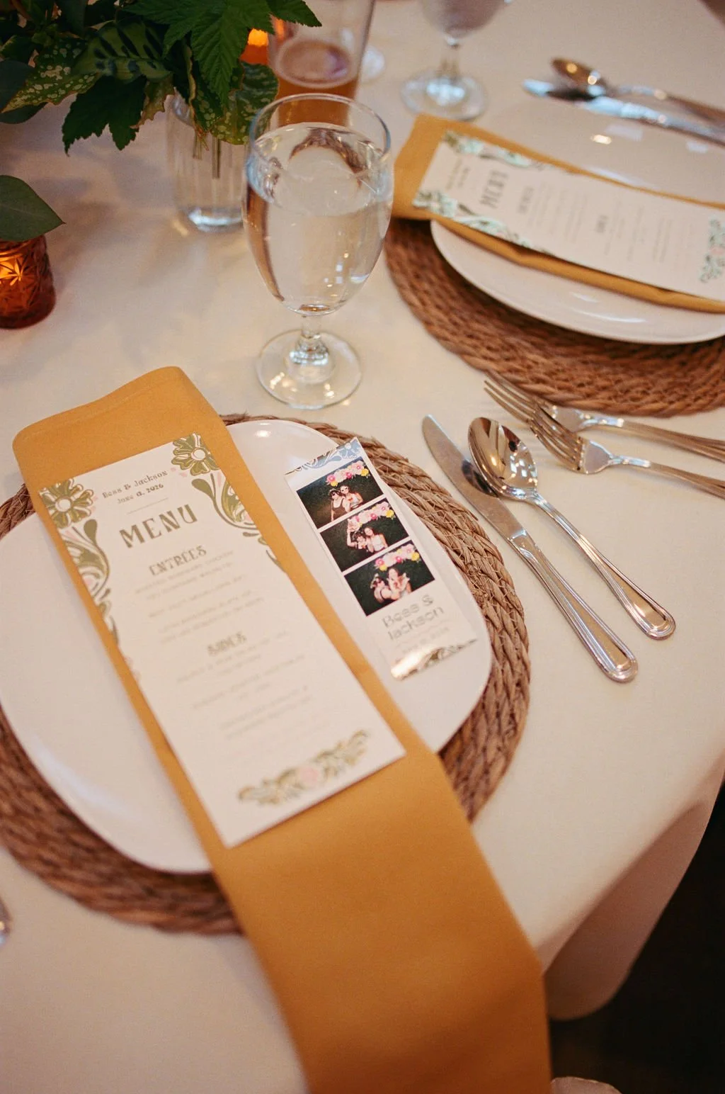 A wedding reception table setup featuring plates with orange napkins, a menu card, a photo booth strip, silverware, a glass of water with ice, a purple drink, and decorative elements including a flower arrangement in a vase and candles.