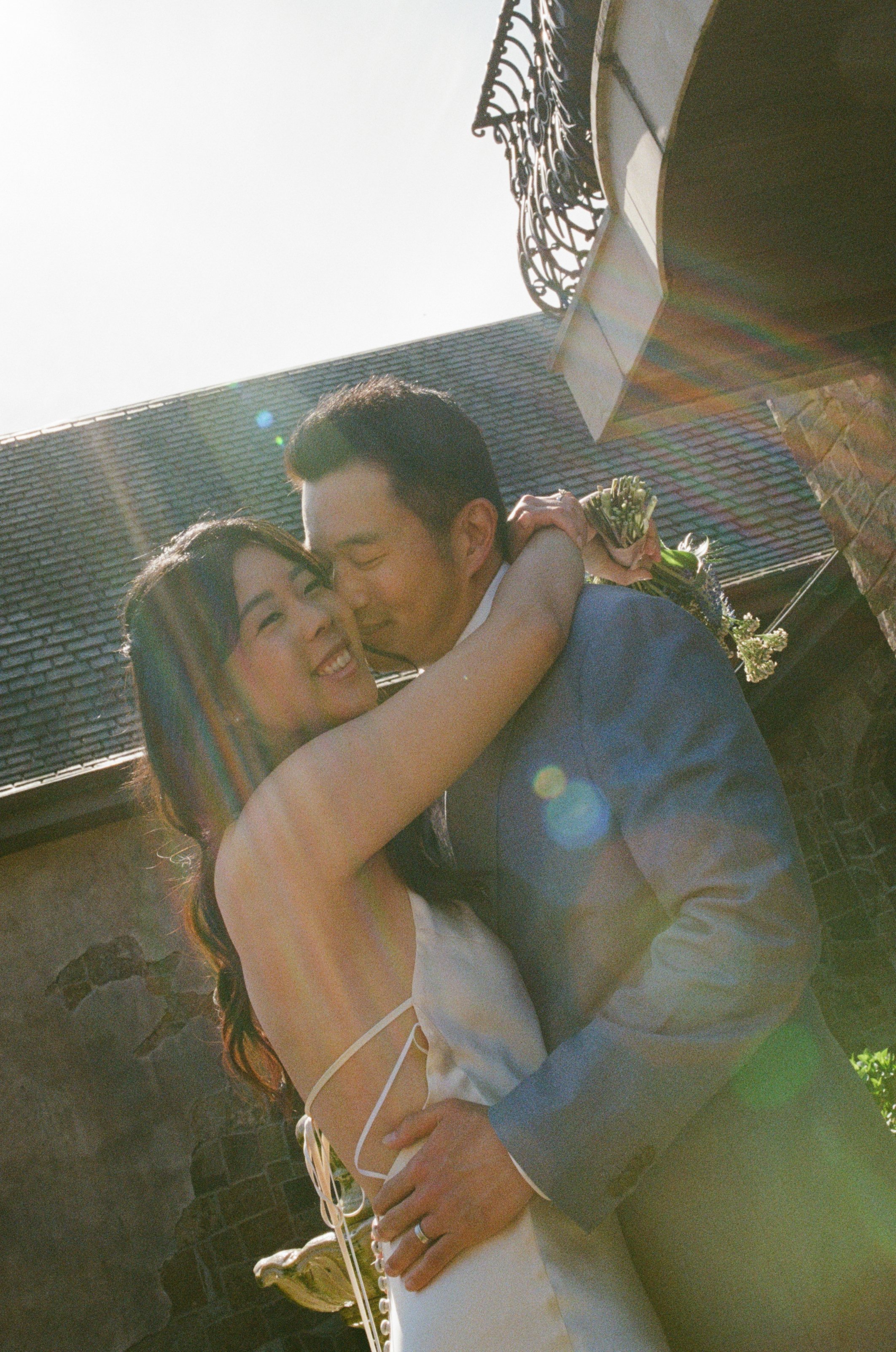 A woman and a man sharing a kiss outdoors during daytime. The woman has long brown hair and is smiling, while the man has short dark hair and is holding her. The scene is lit with sunlight, creating a lens flare effect.