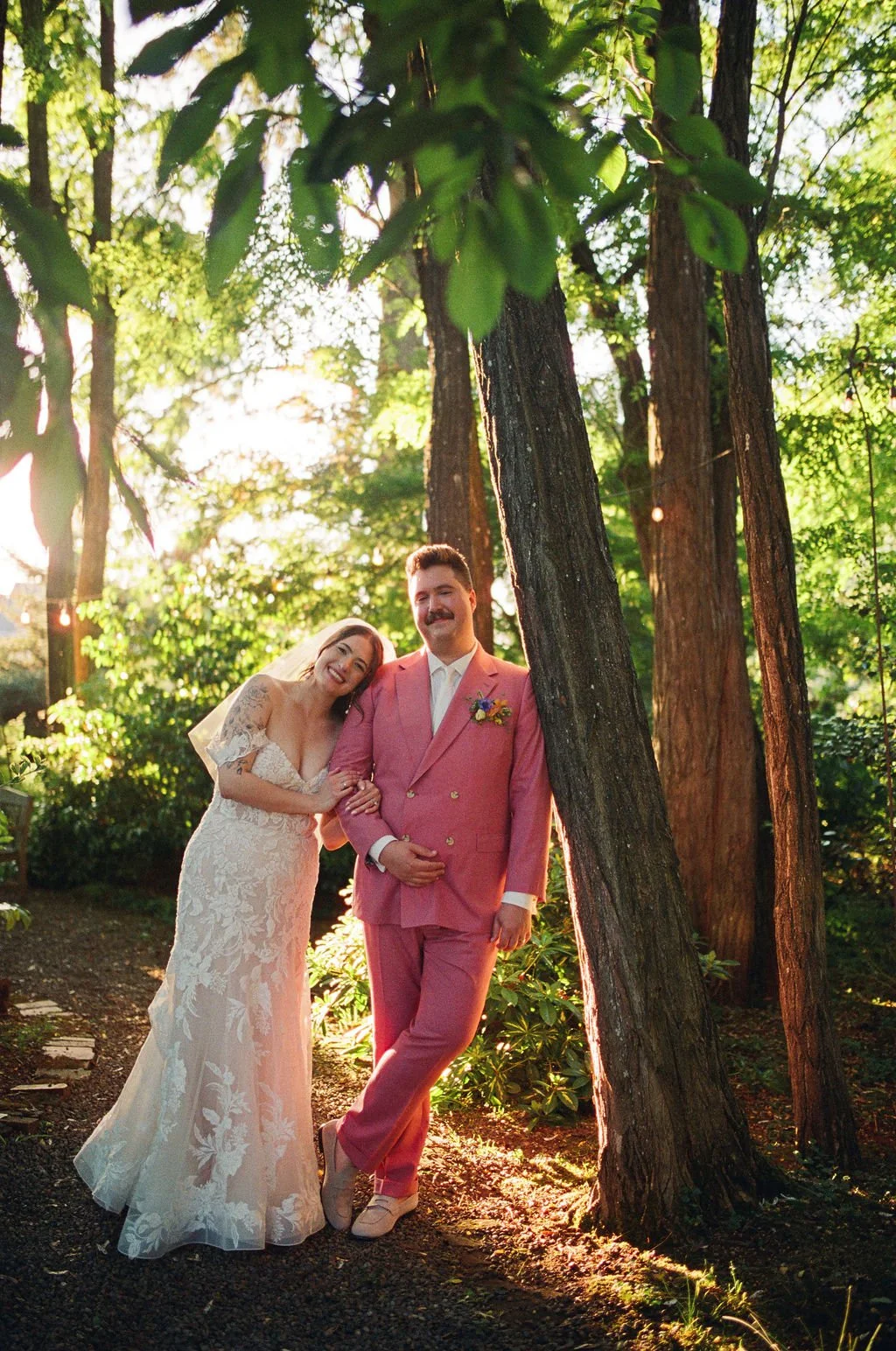 A bride and groom smiling in a forest setting during sunset. The bride wears a lace wedding dress with floral patterns and a veil, leaning affectionately on the groom. The groom wears a pink double-breasted suit with a boutonnière. Sunlight filters t