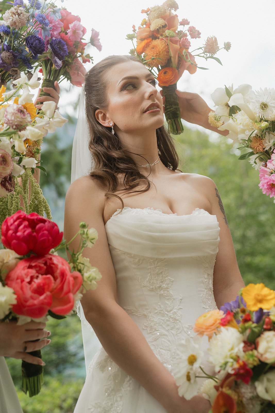 Bride in a strapless white wedding gown standing outside, surrounded by colorful flower bouquets, with an overcast sky and green trees in the background.
