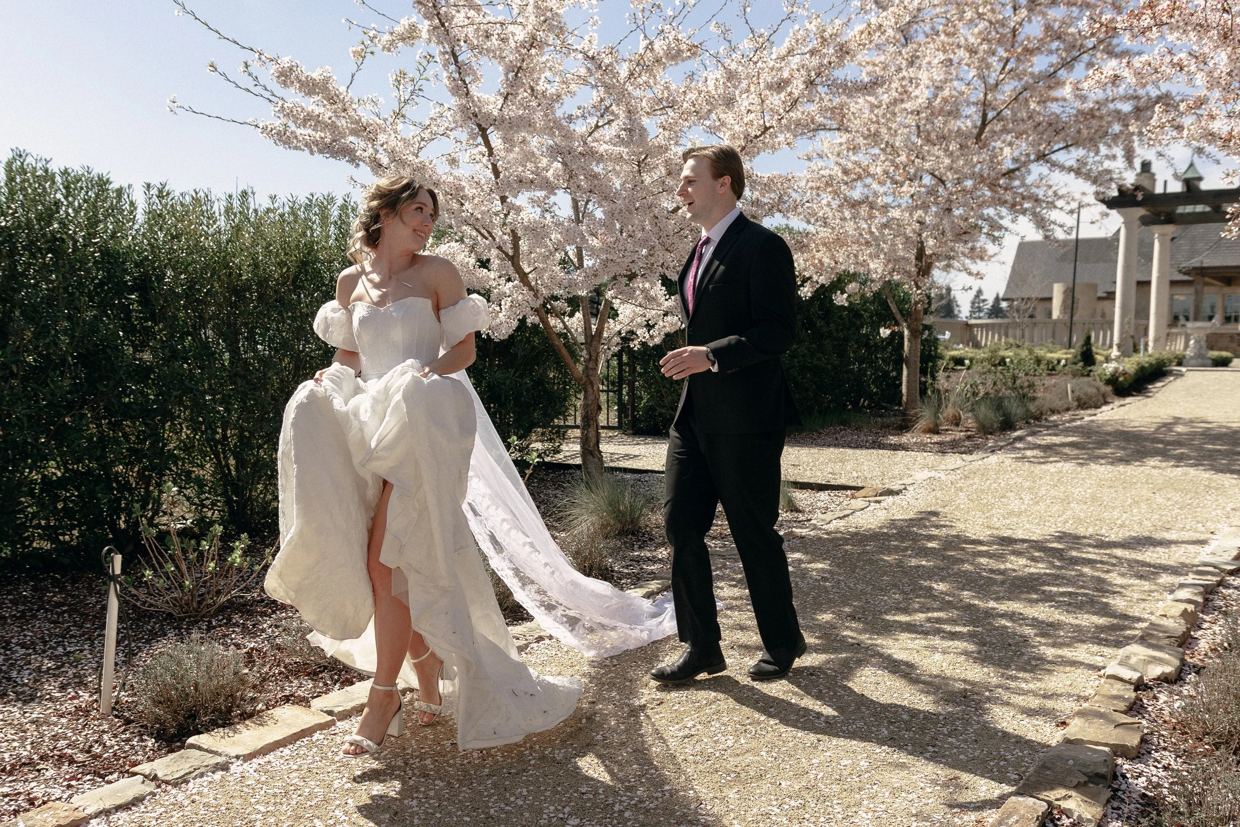 A bride and groom stand outdoors under blooming cherry blossom trees, smiling and looking at each other. The bride is wearing a white strapless wedding gown, and the groom is dressed in a black suit and tie. The setting appears to be a garden or back