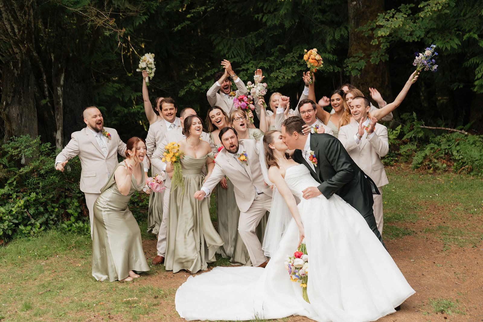 A wedding party of mostly young adults, dressed in formal attire, celebrating outdoors in front of trees. The bride, in a white wedding dress, and the groom, in a dark suit, are kissing in the front. The bridesmaids and groomsmen are behind them, hol