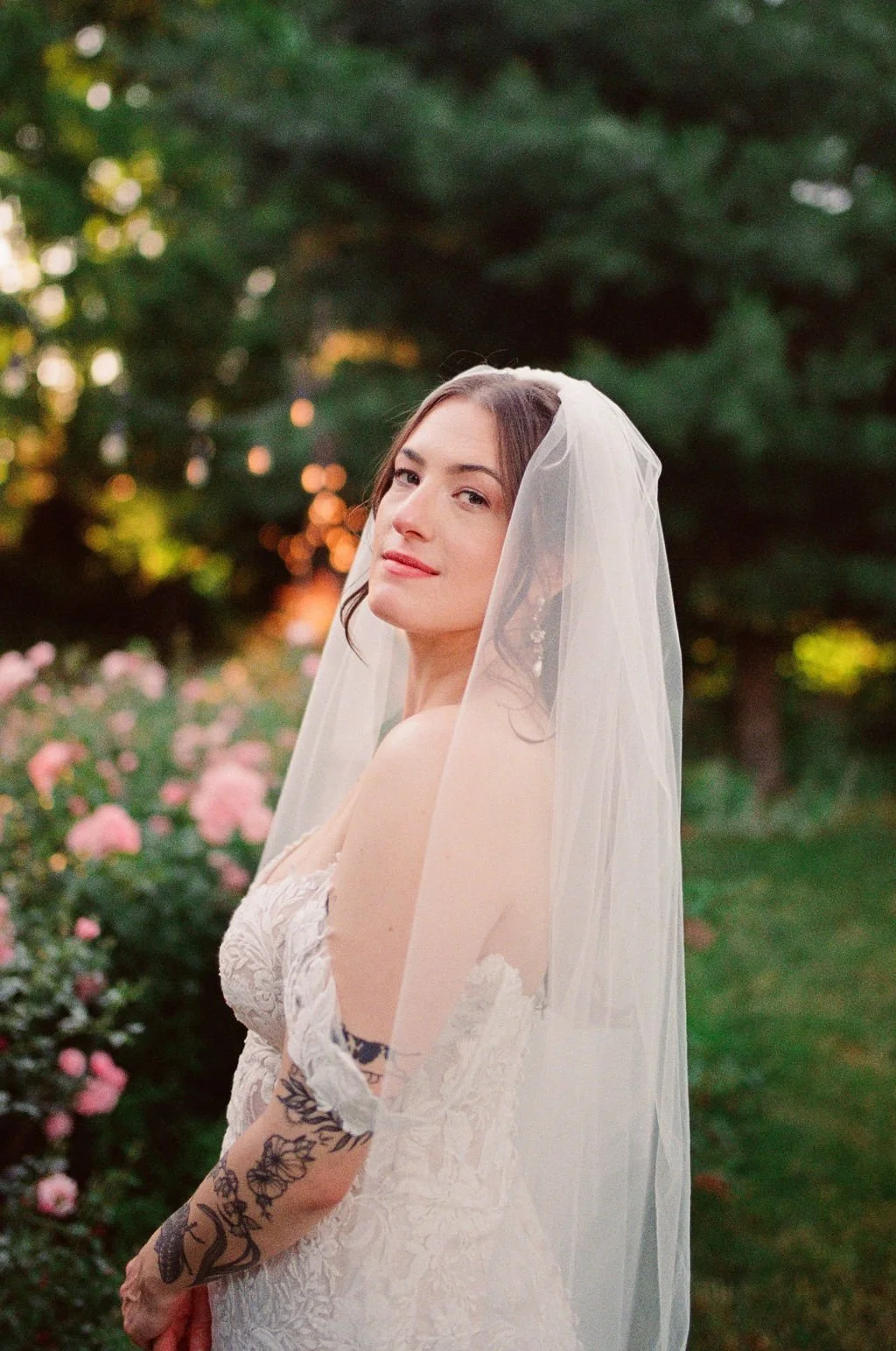 A bride in a wedding dress and veil standing outdoors among pink roses and green trees during sunset.