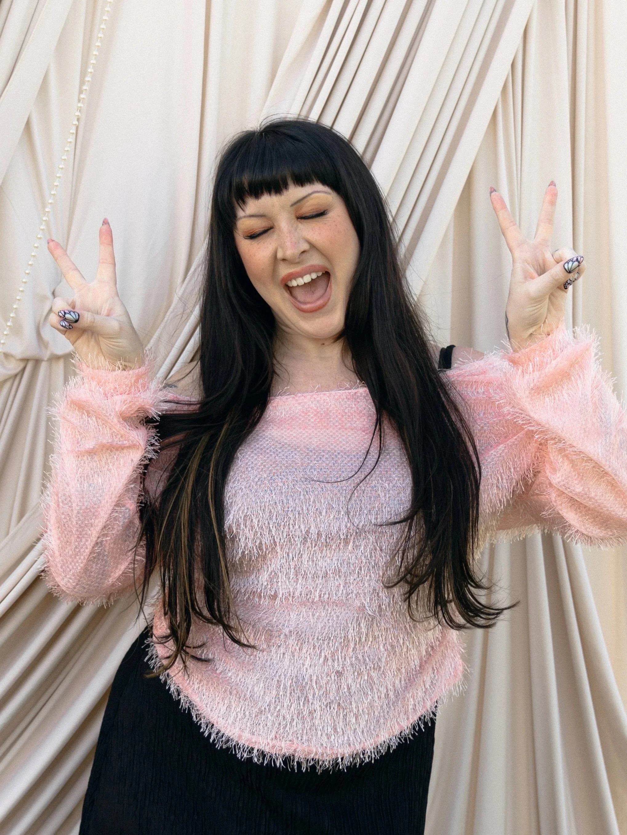 Sloane Simmons, a woman with long black hair and curly bangs, wearing a pink fuzzy sweater, making peace signs with both hands, standing in front of cream-colored curtains.
