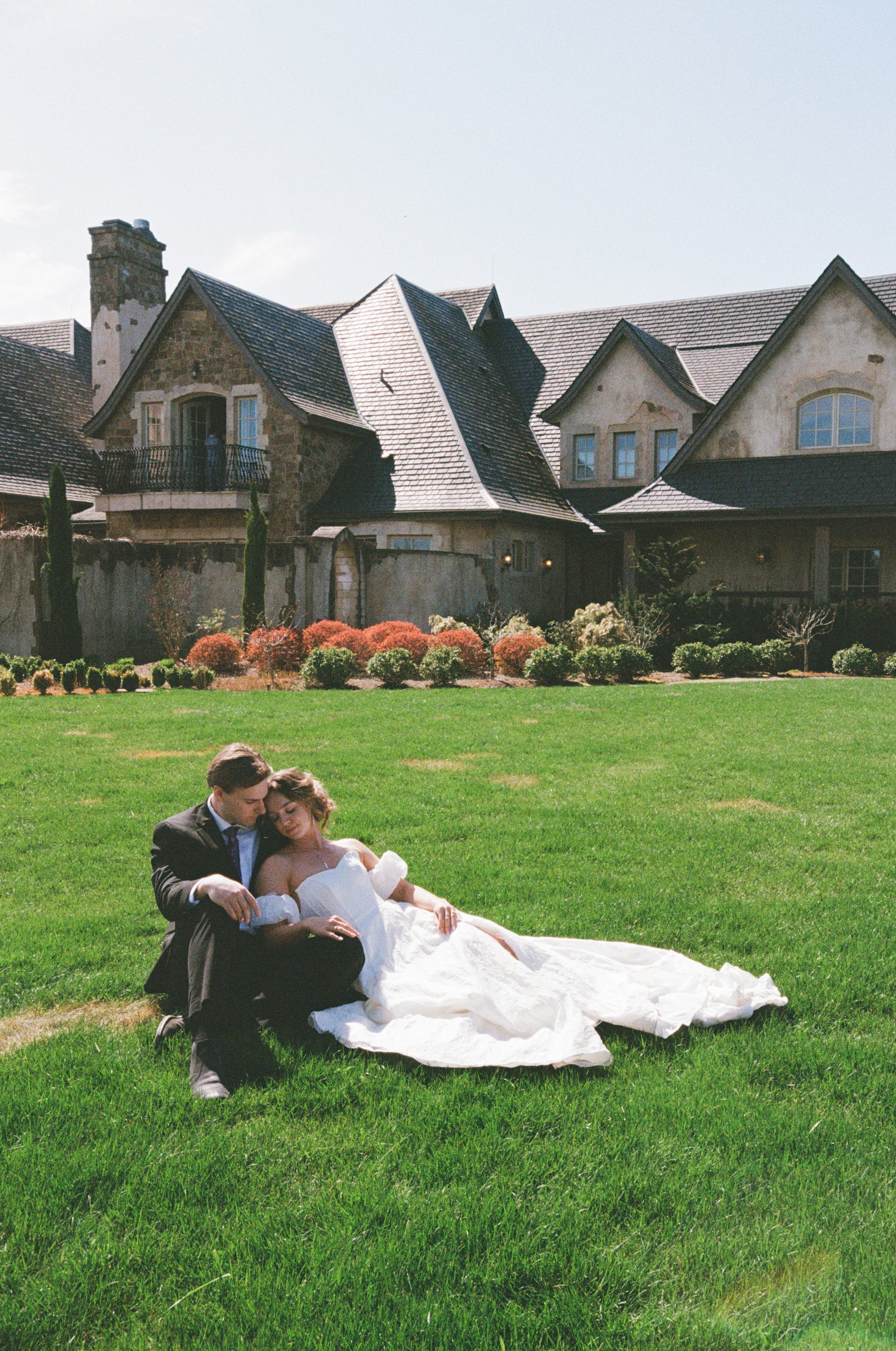 A newlywed couple sitting and leaning on each other on the lush green grass in front of a large house with a slate roof and stone walls, floral bushes and trees in the garden. The groom is wearing a black suit, and the bride is in a white wedding gow
