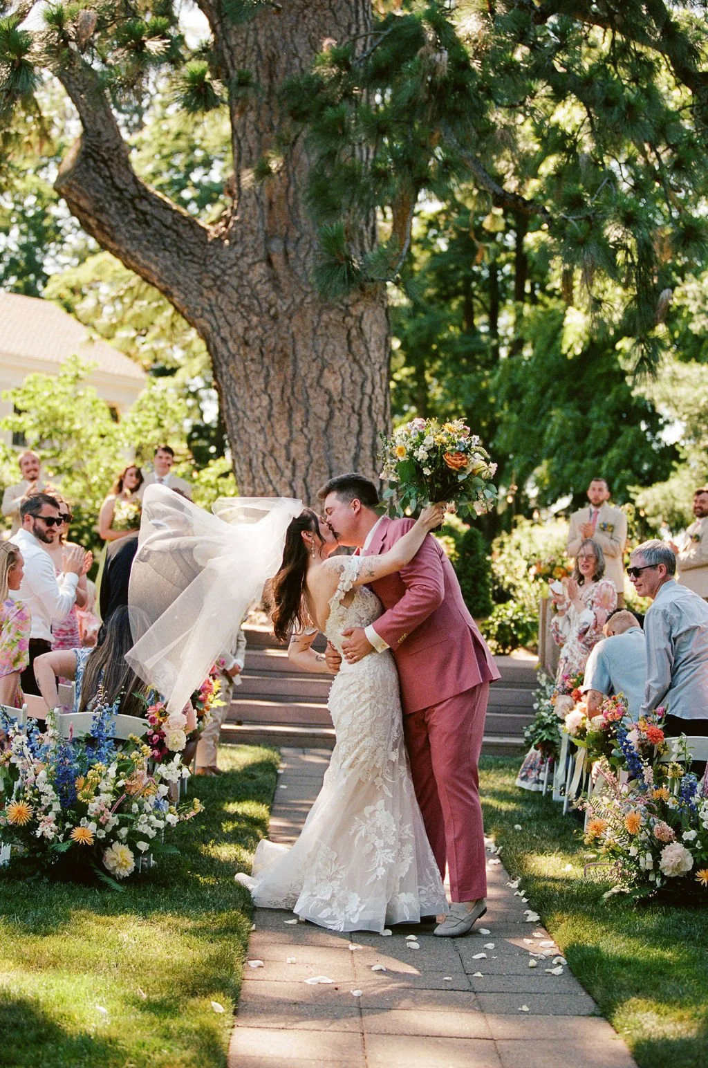 A bride and groom kiss during an outdoor wedding ceremony under a large tree, surrounded by friends and family. The bride holds a bouquet of flowers, and both are dressed in wedding attire.