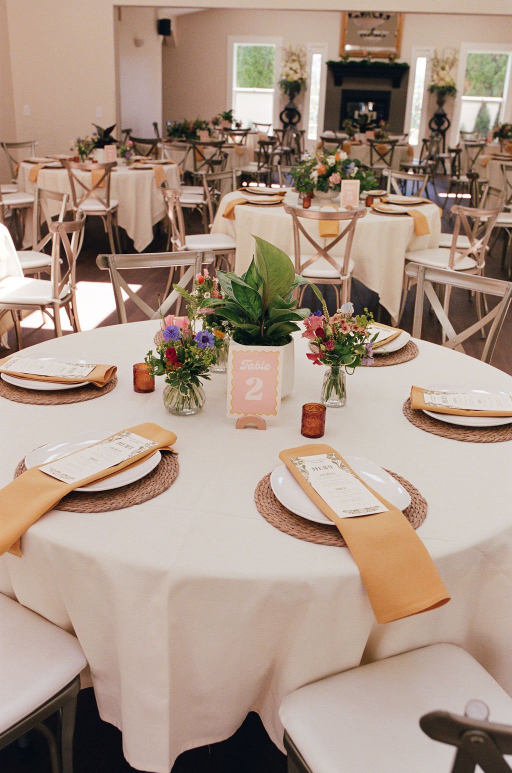 A decorated wedding reception table with floral centerpieces, place settings, menu cards, and a table number sign. The room has multiple round tables, chairs, and a fireplace in the background.