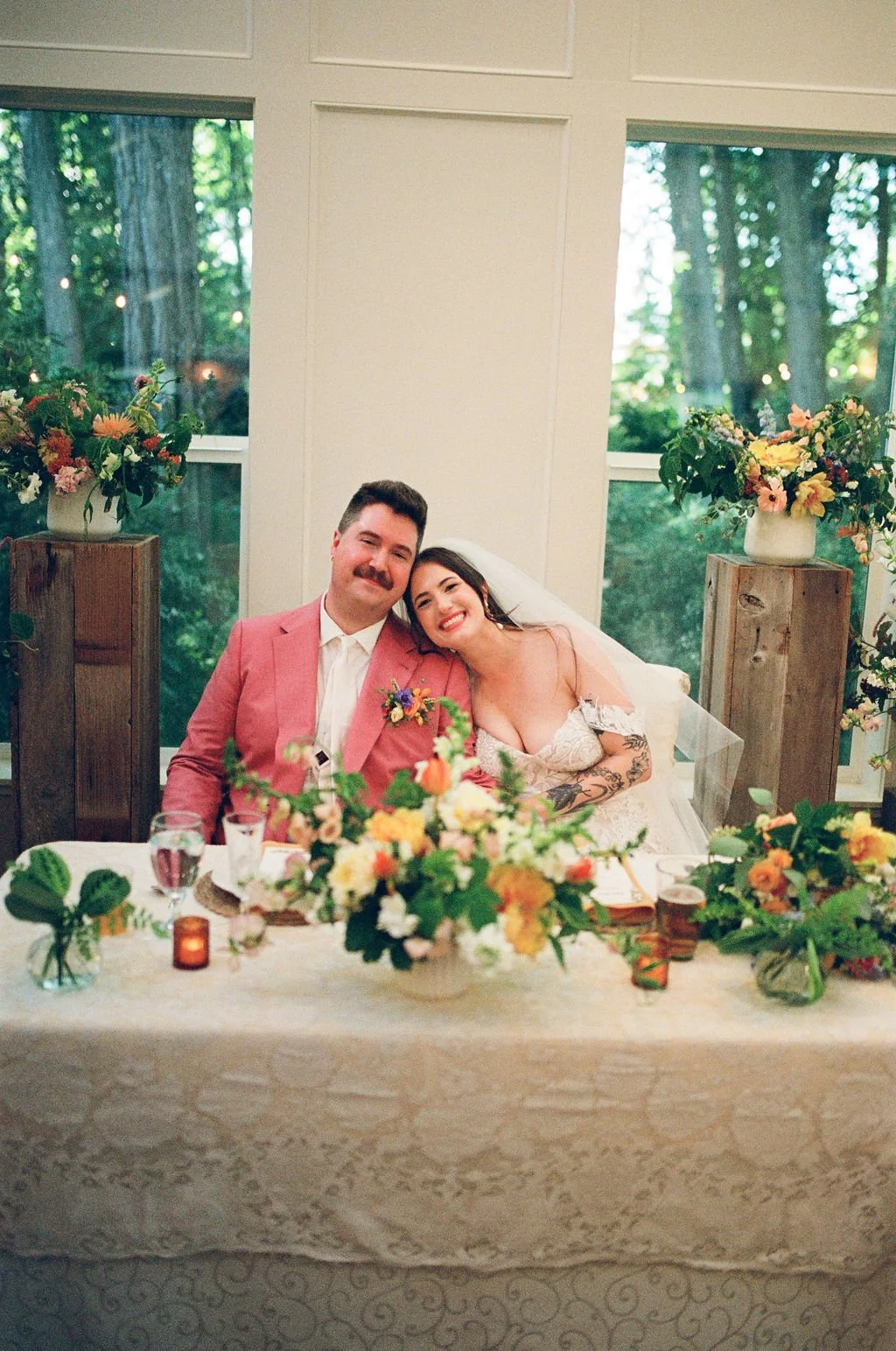 A newlywed couple sitting at a decorated table, smiling with the bride leaning her head on the groom's shoulder. The table has floral arrangements, candles, and glassware, with large flower vases on wooden pedestals behind them, set in a room with la