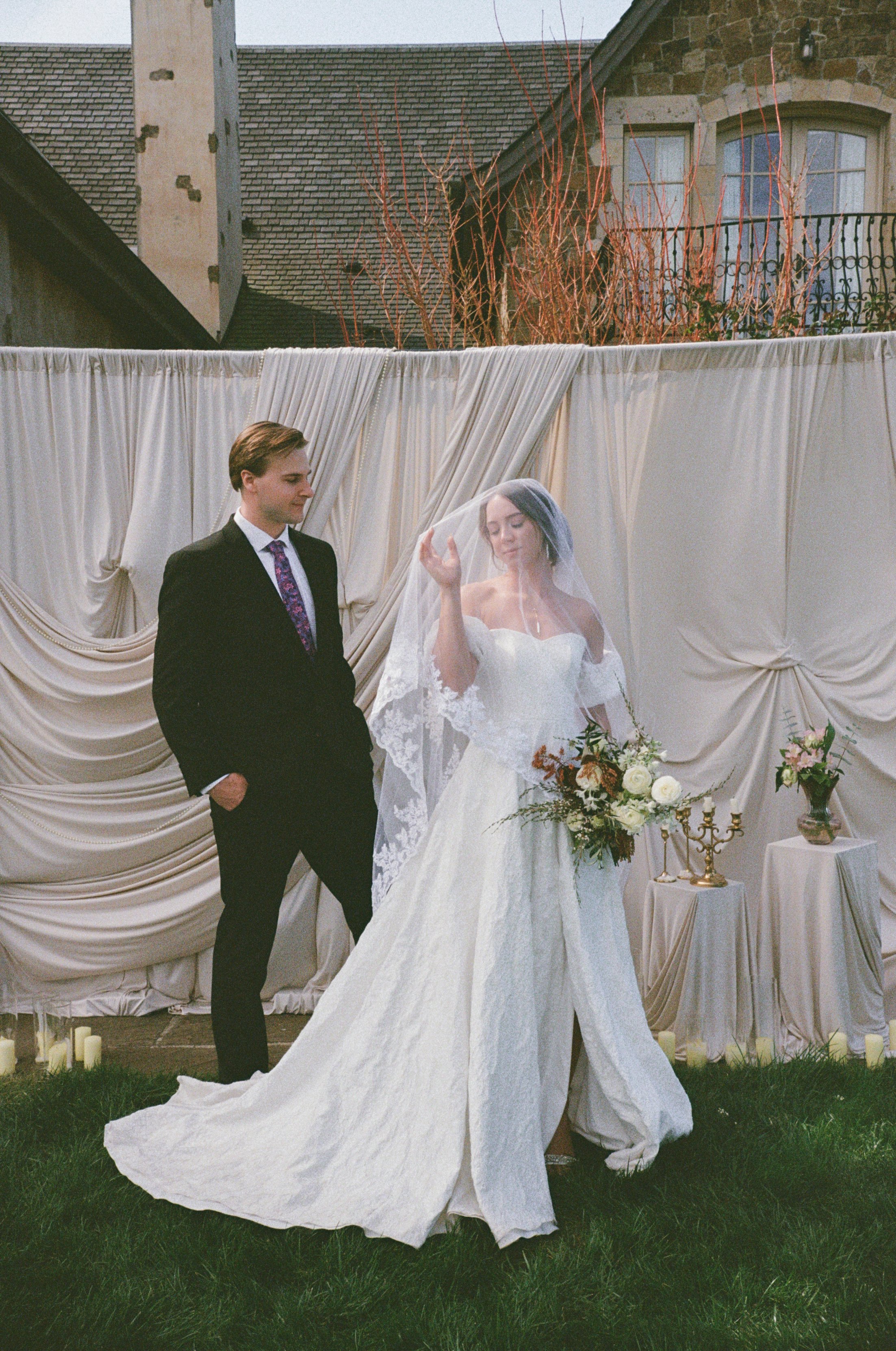 A bride and groom standing in front of a cream-colored curtain backdrop during their outdoor wedding ceremony. The bride wears a white wedding dress with a long train and a veil, and is holding a bouquet of flowers. The groom is dressed in a black su