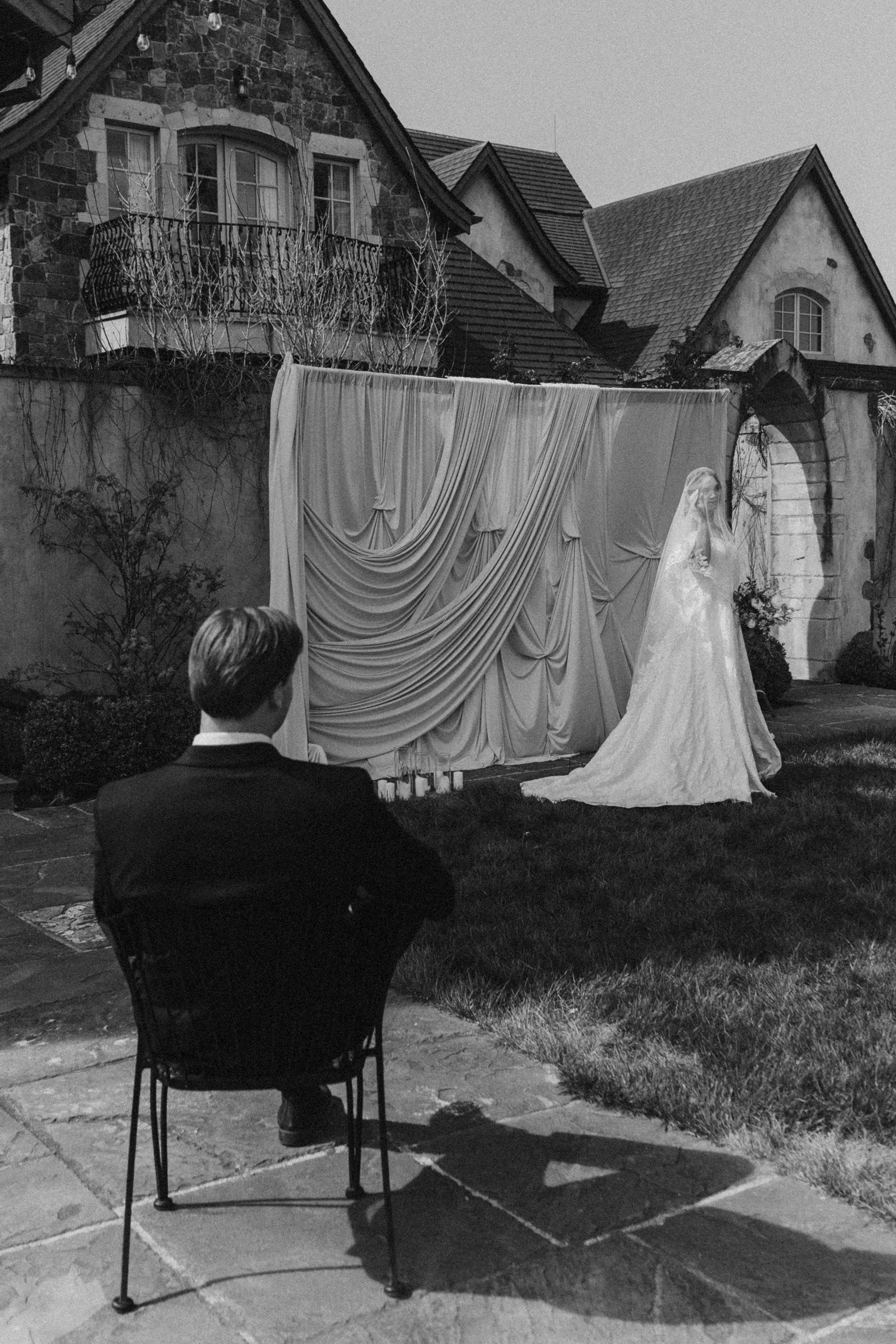 A man in a tuxedo sitting outdoors, observing a wedding ceremony in front of a draped backdrop with a wedding dress displayed to the right. The scene is set in a backyard with a stone house and patio area, with the ceremony taking place during the ev