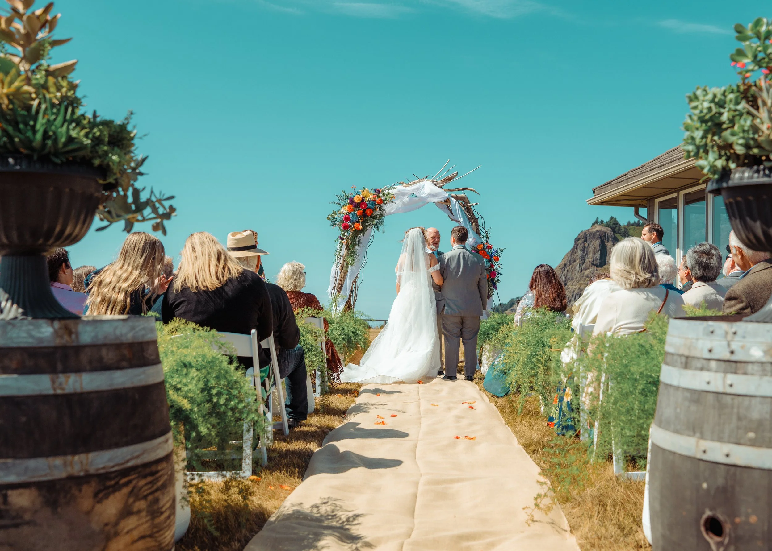 A wedding ceremony outdoors with a bride and groom standing under a floral arch, surrounded by seated guests, with large potted plants in the foreground and rocky hills in the background.