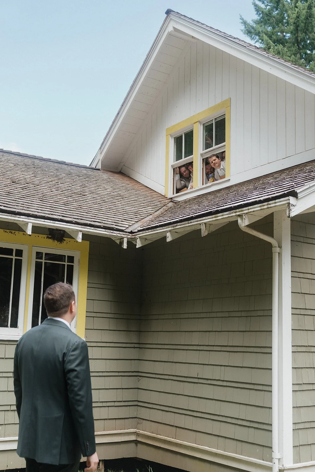 A man in a suit standing outside, looking up at children looking out from a second-story window of a house.