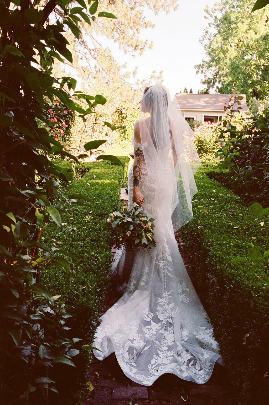 A bride in a white lace wedding dress and veil standing on a garden path, holding a bouquet, with greenery and a small house in the background.