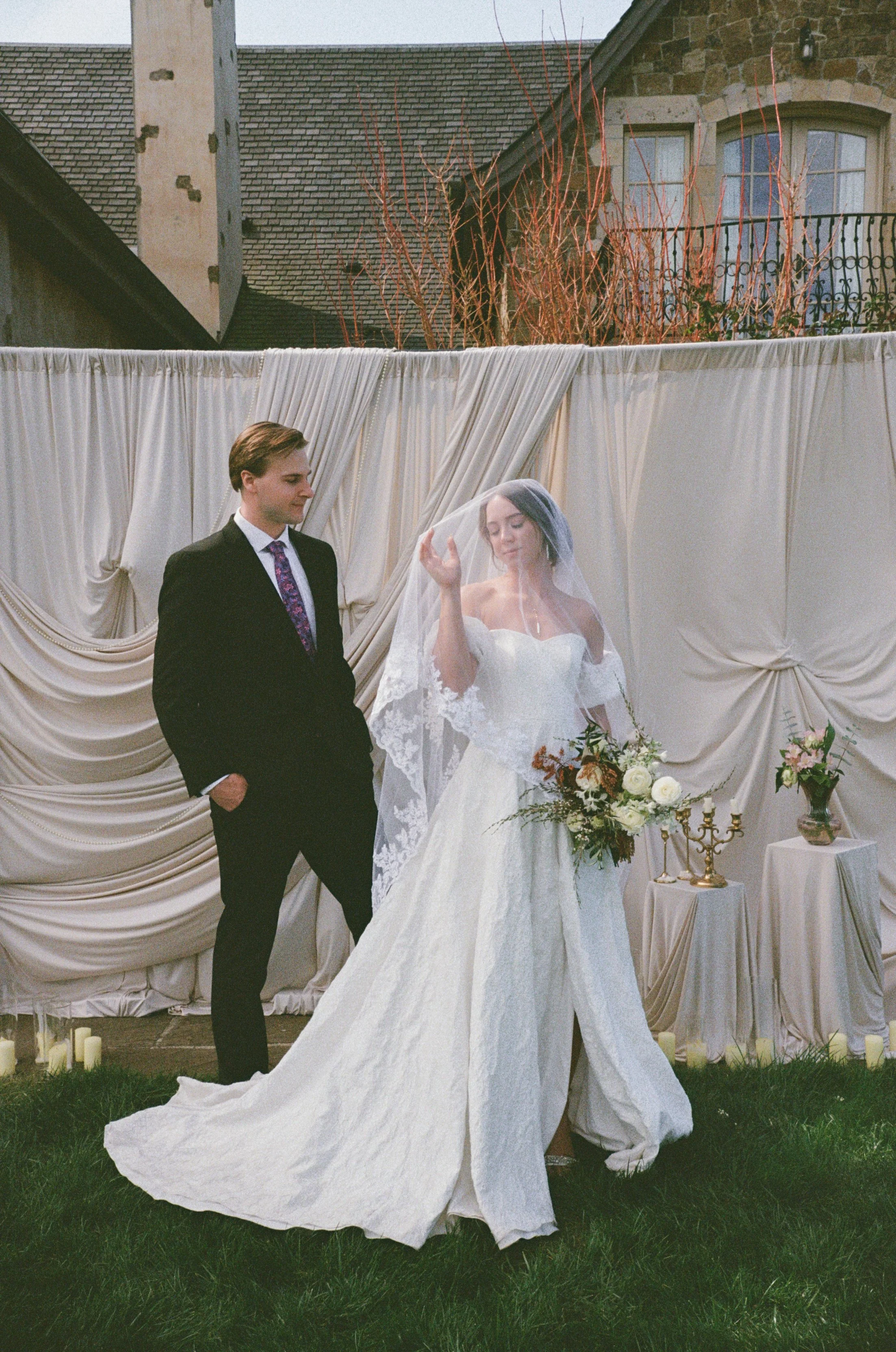 A bride and groom standing in front of a white draped backdrop during a wedding ceremony outside. The bride has blue hair and is wearing a white strapless wedding gown and veil, holding a bouquet of white roses and greenery. The groom has light brown hair and is wearing a black suit with a white shirt and a colorful purple and pink patterned tie.