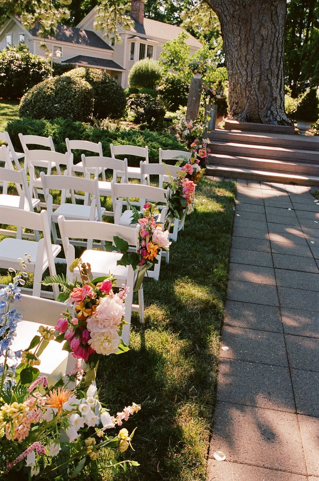 Outdoor wedding ceremony setup with white chairs and floral decorations along a rope, with a house and large tree in the background.
