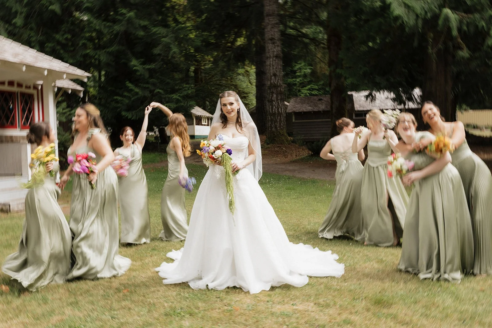A bride in a white wedding gown holding a bouquet of colorful flowers, standing on grass in a wooded outdoor setting with bridesmaids dancing and laughing around her.