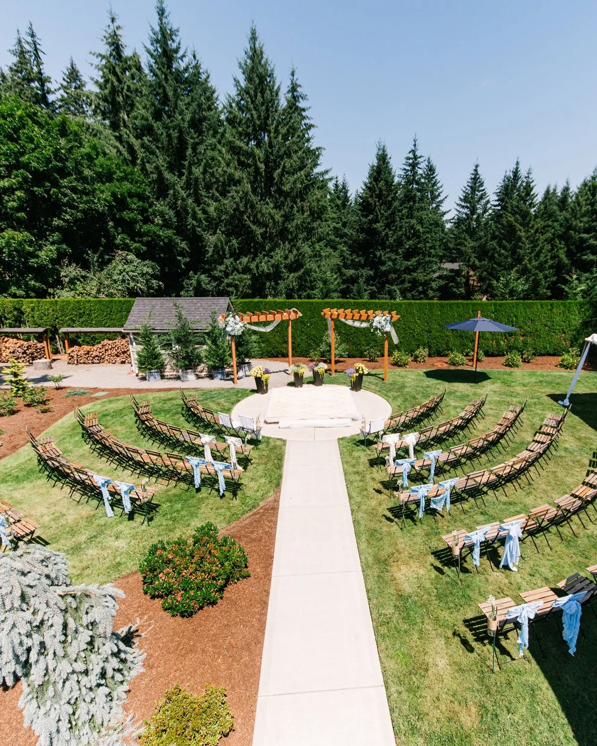 Outdoor wedding ceremony setup in a garden with rows of wooden chairs decorated with white and blue ribbons, an aisle, and a small stage with floral arrangements, surrounded by green trees and a neatly trimmed hedge.