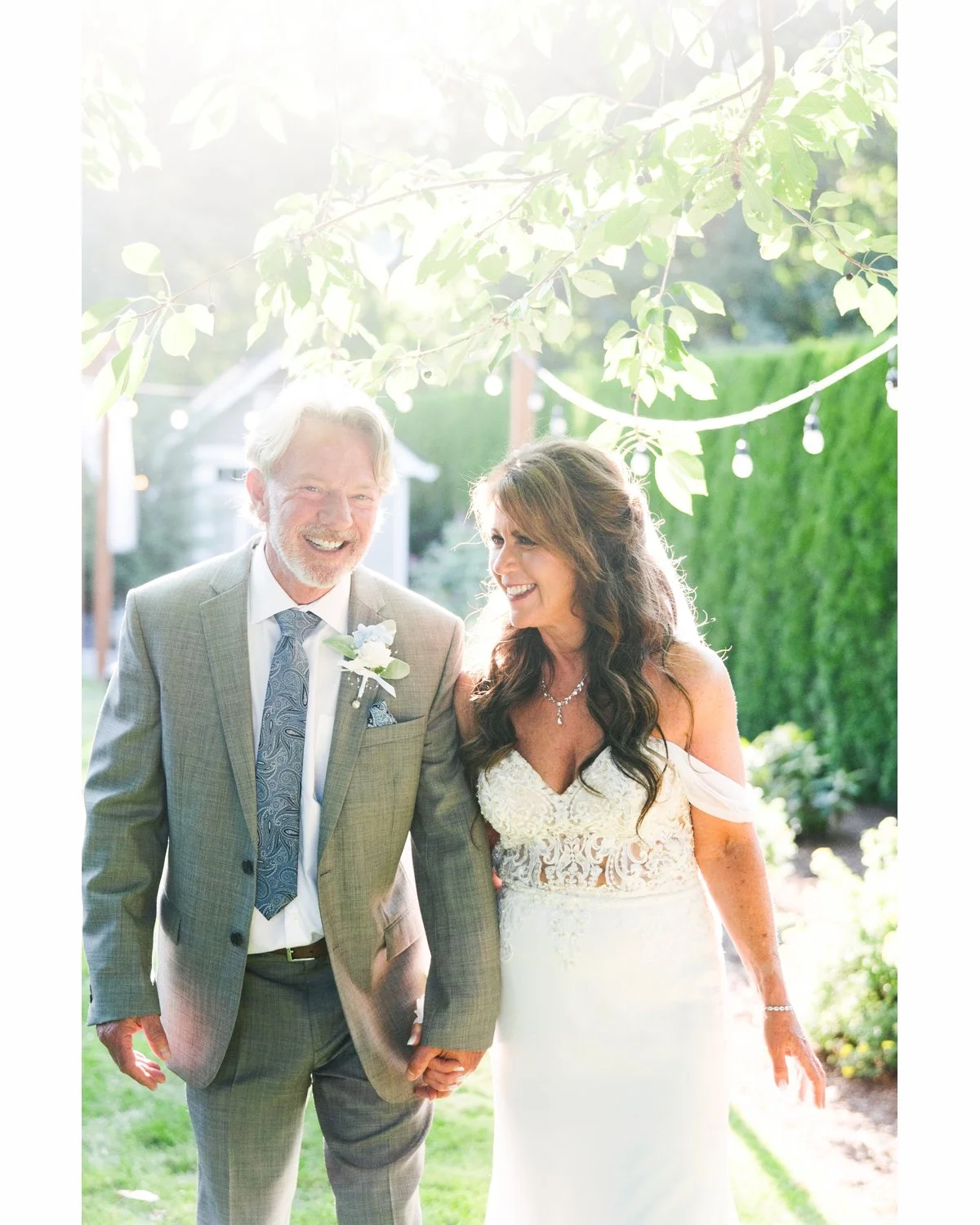A joyful couple, a groom and bride, walking hand in hand outdoors during a wedding celebration, smiling and looking happy with sunlight filtering through tree branches.