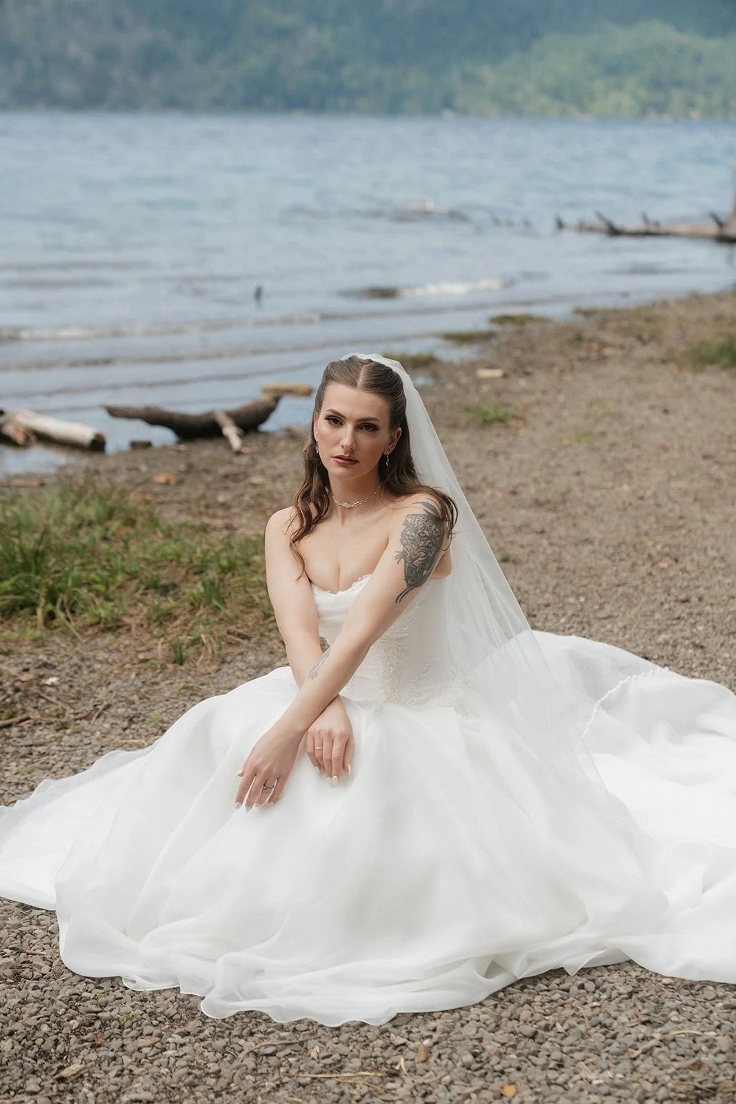 A woman in a white wedding dress and veil sitting on a rocky beach near a lake, with forested hills in the background.