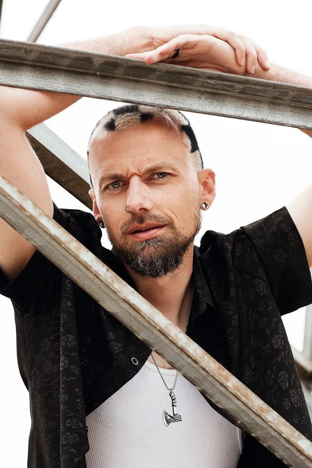 Man with a beard, tattoos, earrings, and a necklace with a hammer pendant, leaning on a metal ladder, looking at the camera.