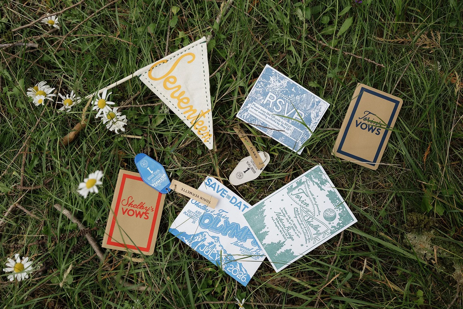 Various wedding invitations, RSVP cards, and event flags placed on grass with small white daisies.