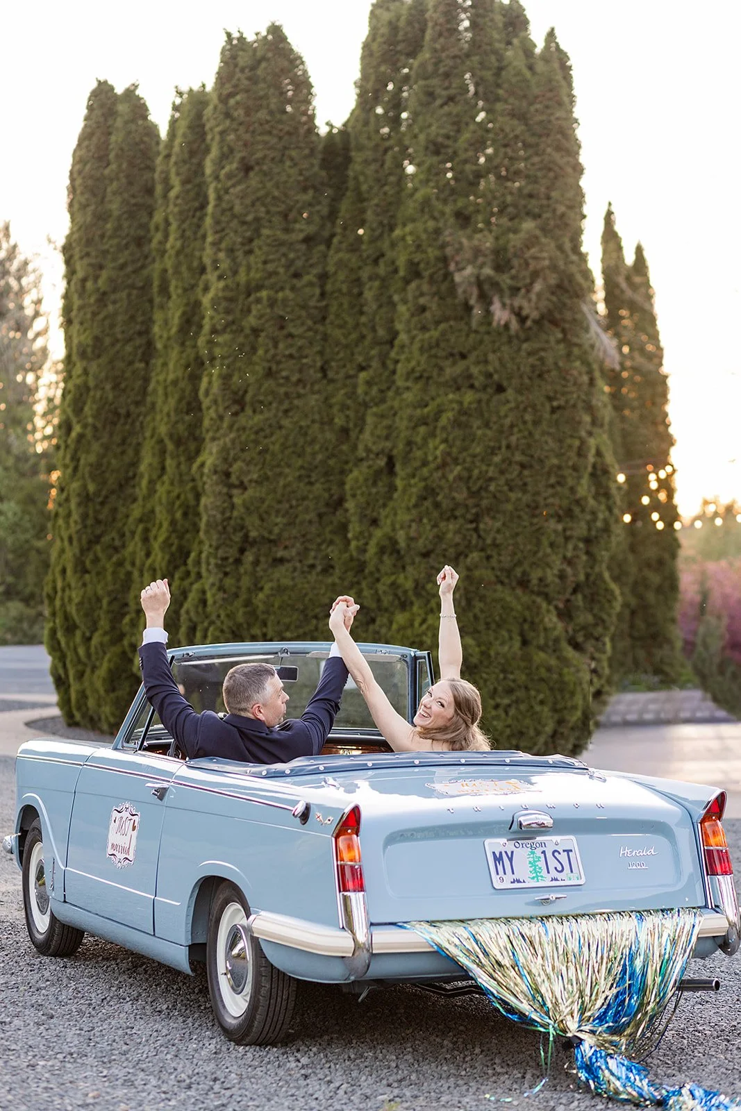 Two young people celebrating in a vintage convertible car decorated for a special occasion, with a large green hedge in the background and a shiny decoration at the back of the car.