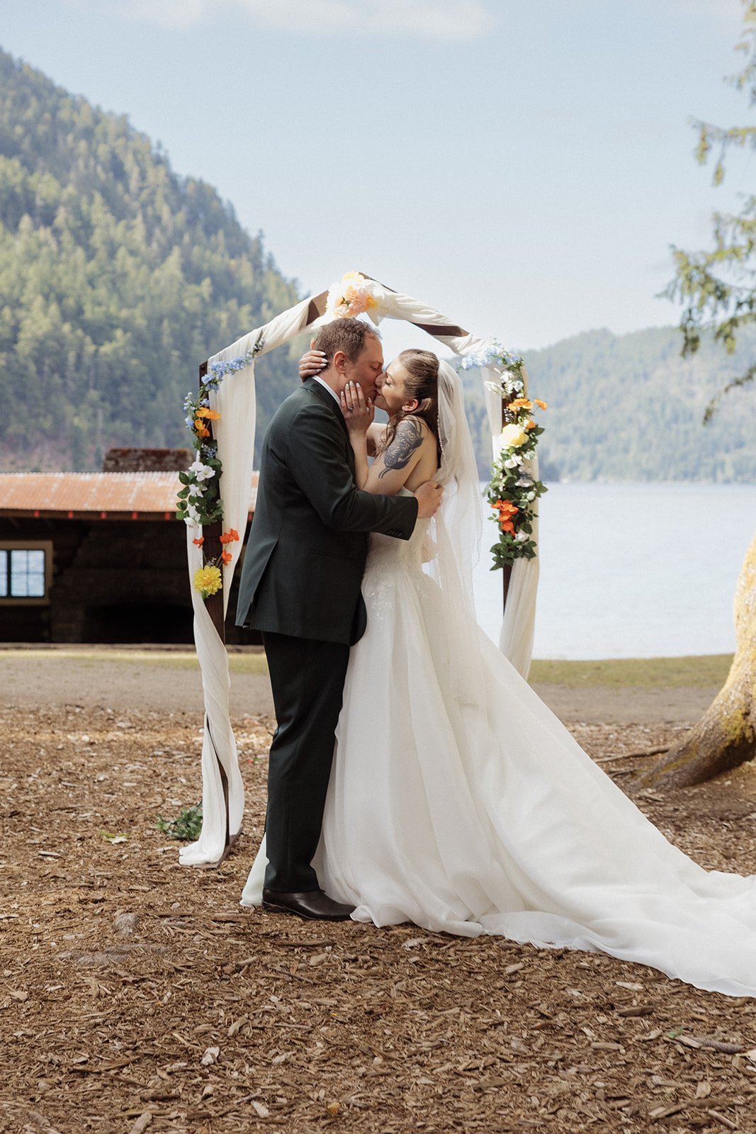 A couple in wedding attire sharing a kiss under a floral wedding arch outdoors, with trees, water, and mountains in the background.