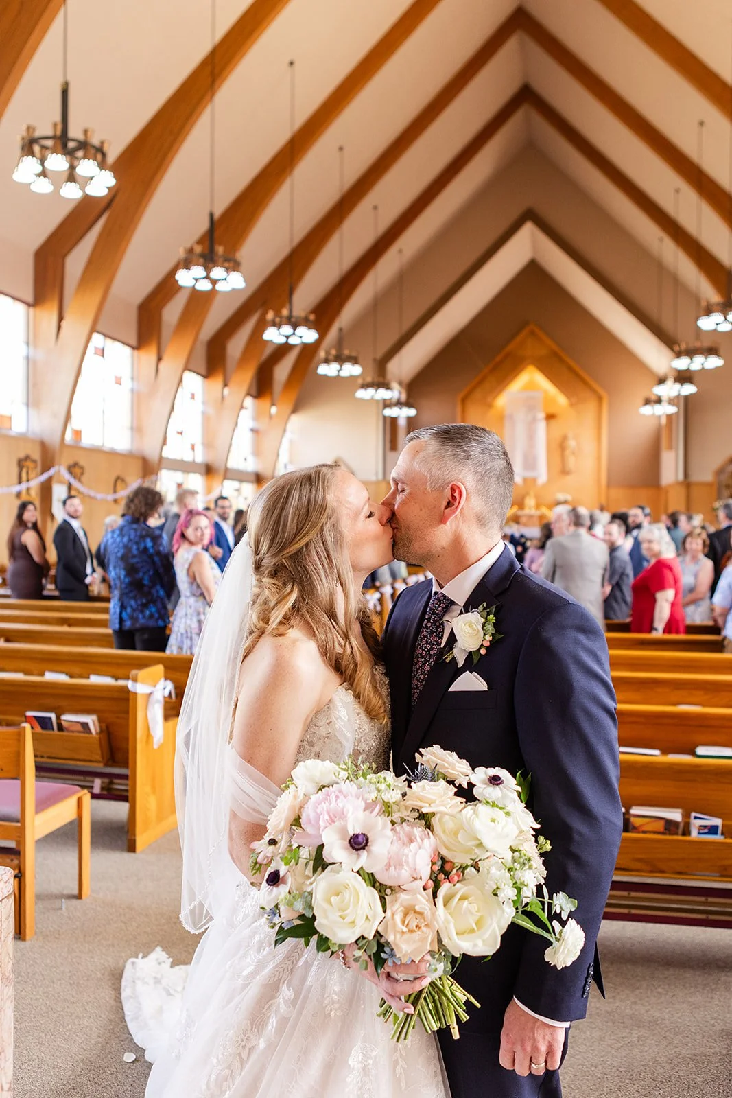 A bride and groom kiss in a church, with wedding guests in the background, during a wedding ceremony. The bride holds a bouquet of flowers, and the church features wooden architecture with chandeliers.