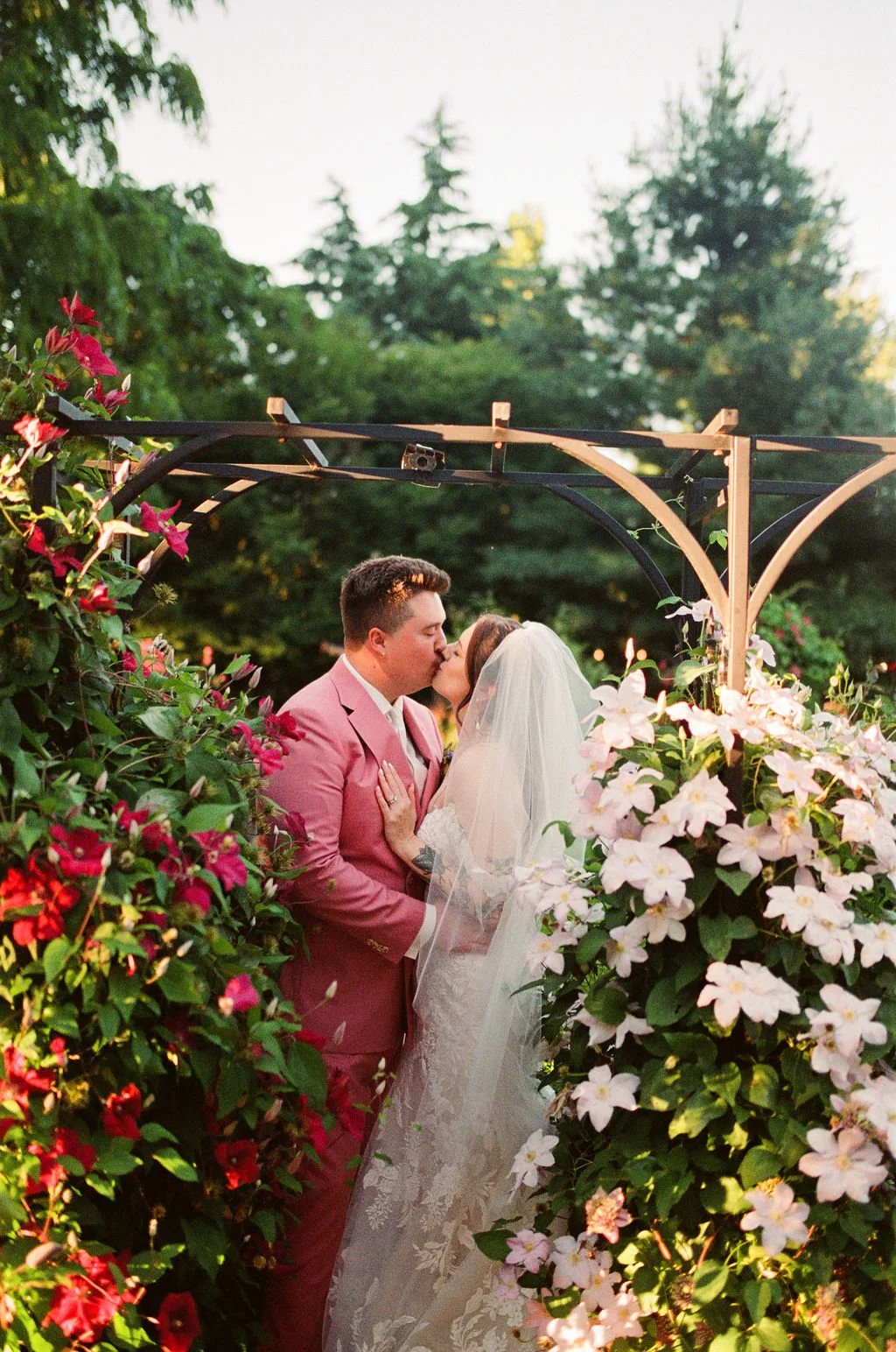 A newlywed couple sharing a kiss under a floral archway, surrounded by pink and white flowers, with green trees in the background during sunset.