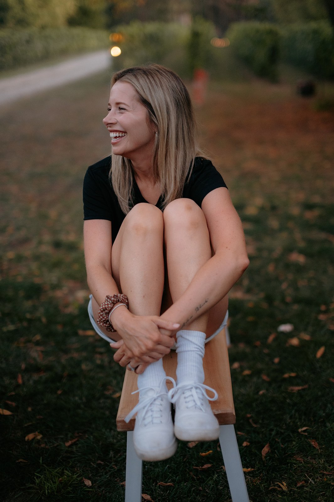 A woman sitting on a wooden stool outdoors, smiling and laughing, with her knees pulled to her chest, wearing a black t-shirt, white shorts, white sneakers, and white socks, in a natural setting during dusk.