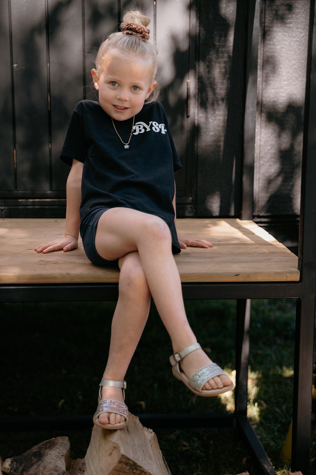 A young girl with blonde hair in a bun, wearing a black t-shirt, black shorts, and glittery sandals, sitting on a wooden platform outdoors with a dark metal fence behind her.