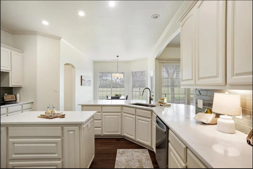 Bright kitchen with white cabinets, countertop, and a dining area in the background, illuminated by natural light through multiple windows.