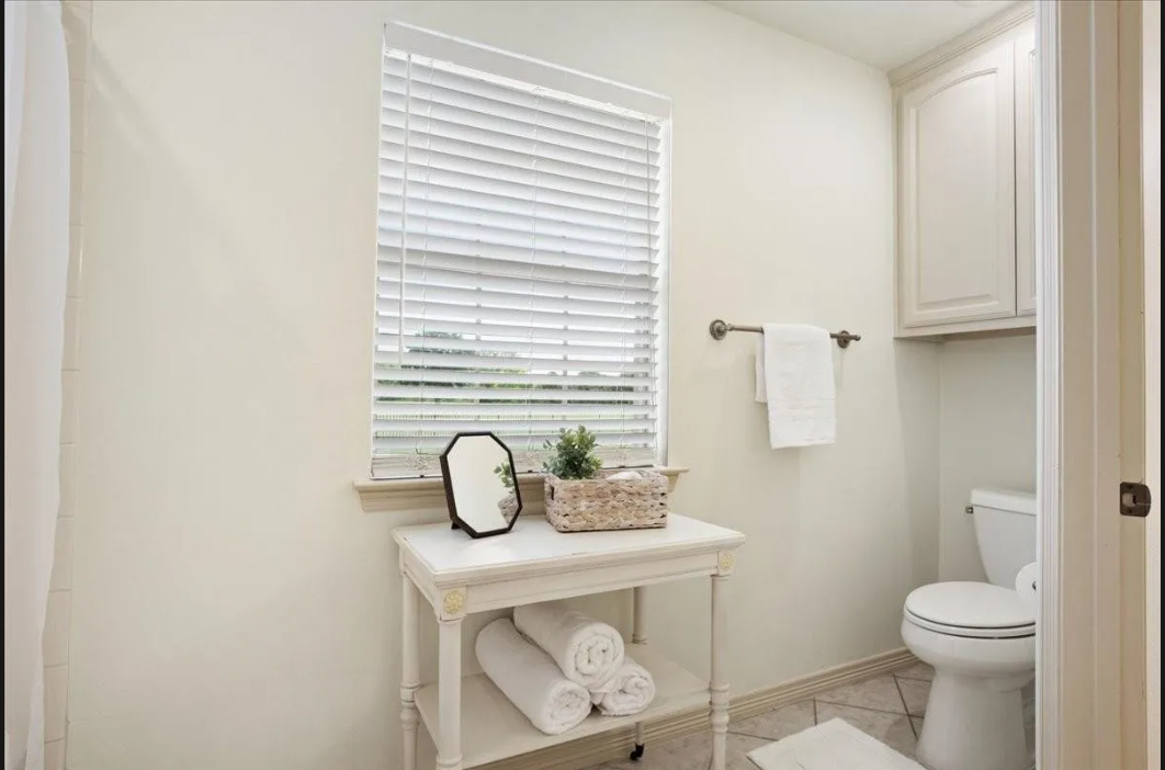 A clean, bright bathroom with a small white table holding towels and a plant, a window with blinds, and a toilet.