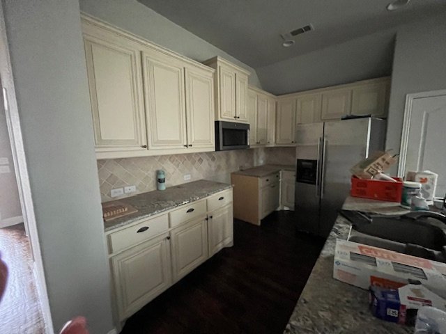 A kitchen with light-colored cabinets, granite countertops, a stainless steel refrigerator, and a built-in microwave. The countertops have various items, including a red container and boxes.